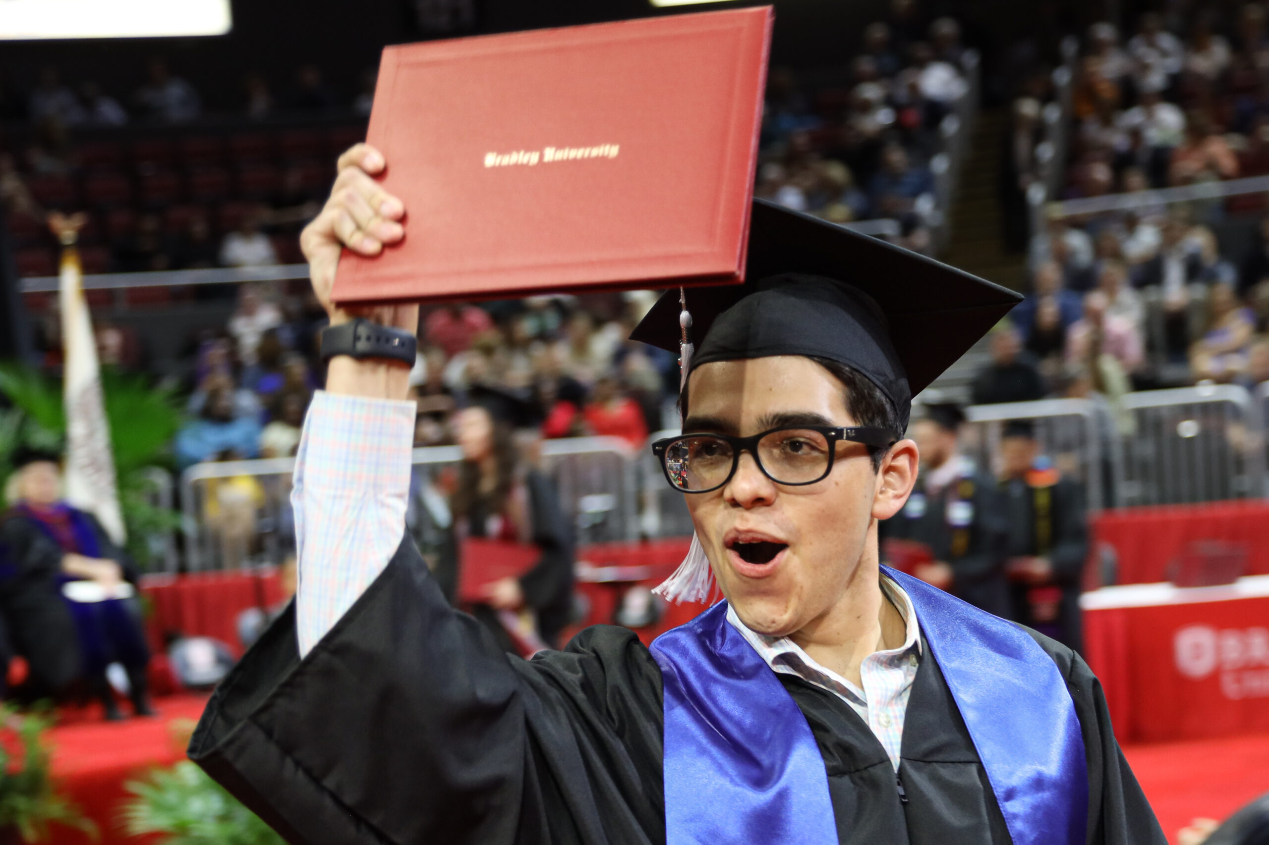 A male in a graduation gown at hat holding up his diploma at a graduation ceremony.