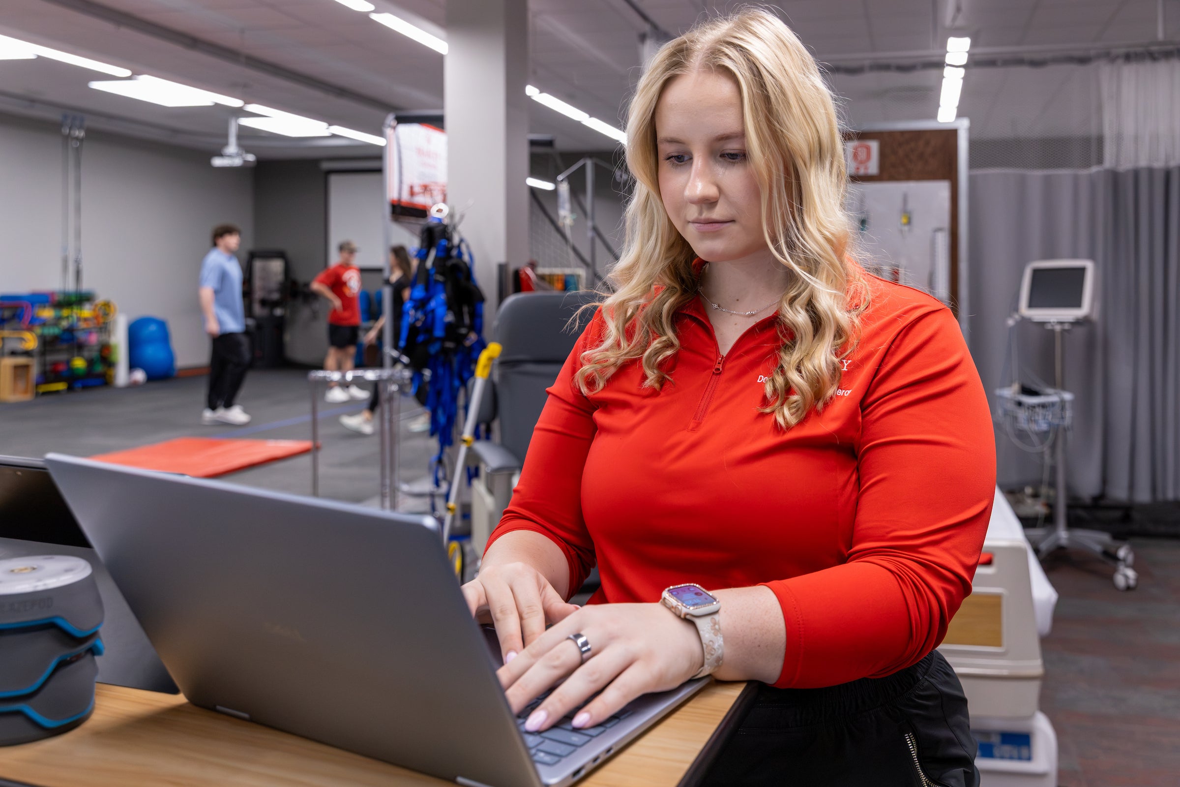 A student clinician working on a laptop while therapy sessions happen behind her