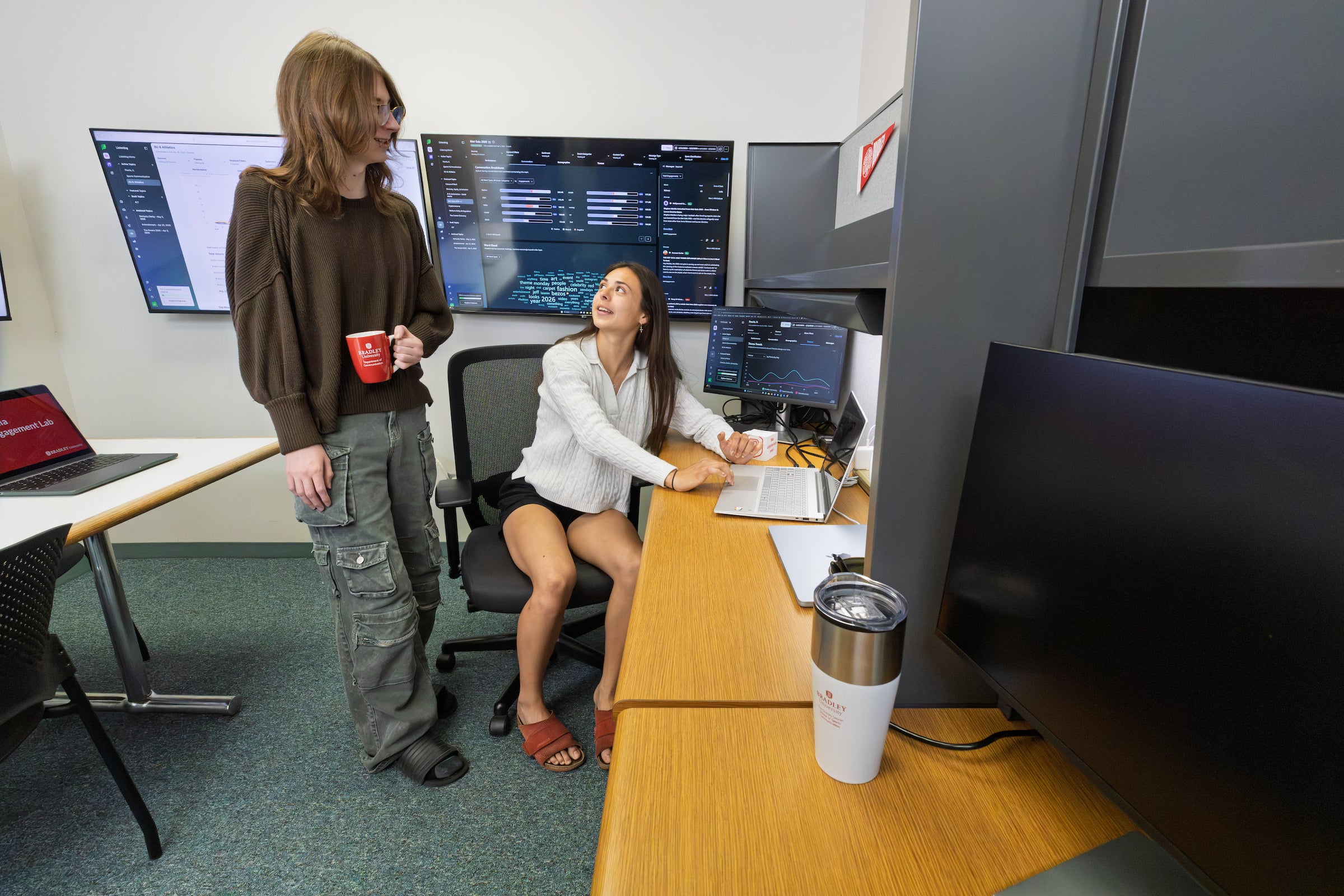 Two female students having a discussion at a table with analytical information on a screen behind them