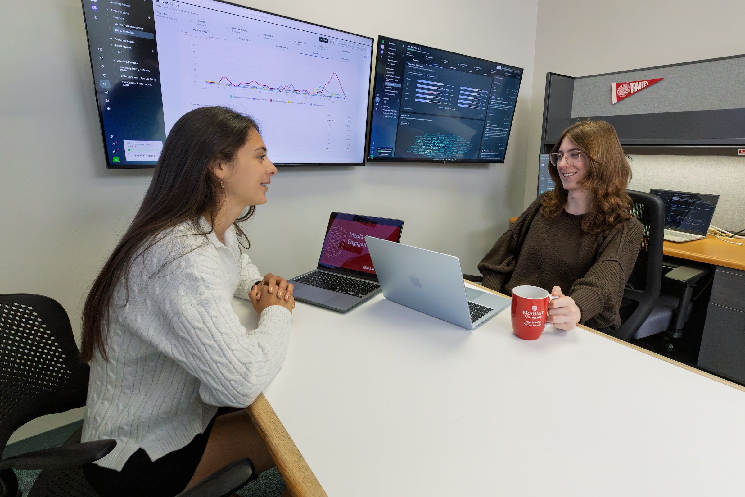 Two female students having a discussion at a table with analytical information on a screen behind them