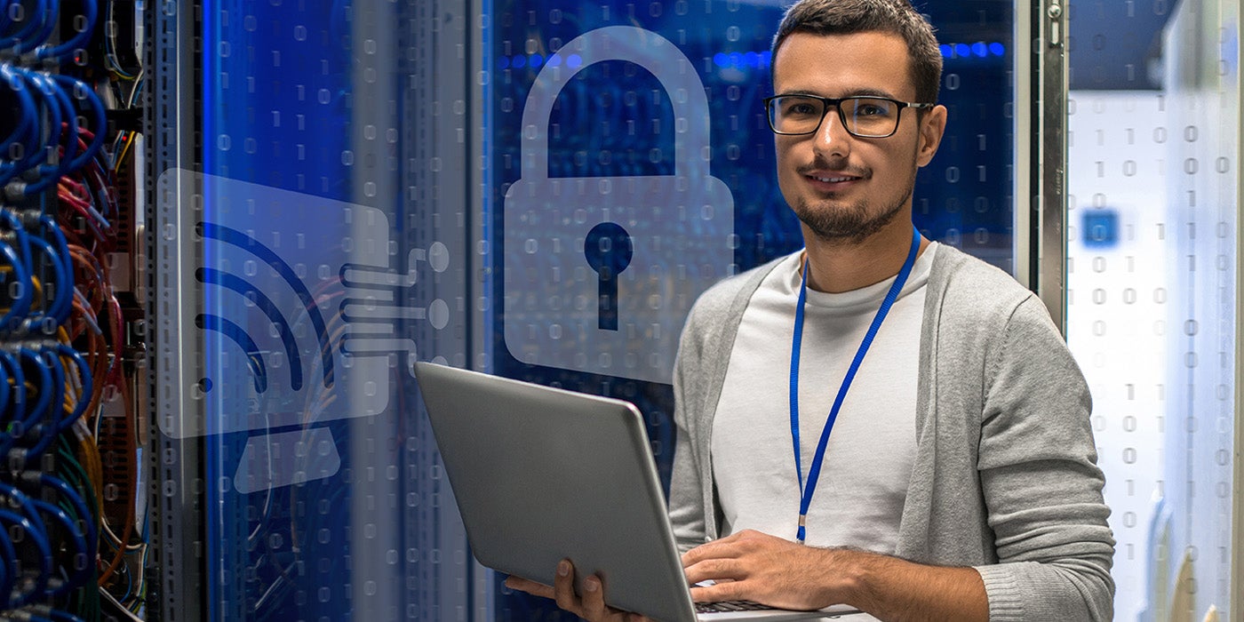 A man standing in a server room holding a laptop while looking a the camera