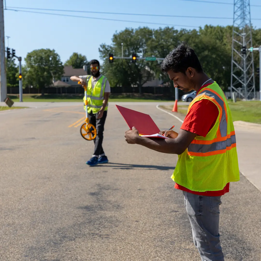 Two students with reflective vests and survey equipment making notes on a clipboard