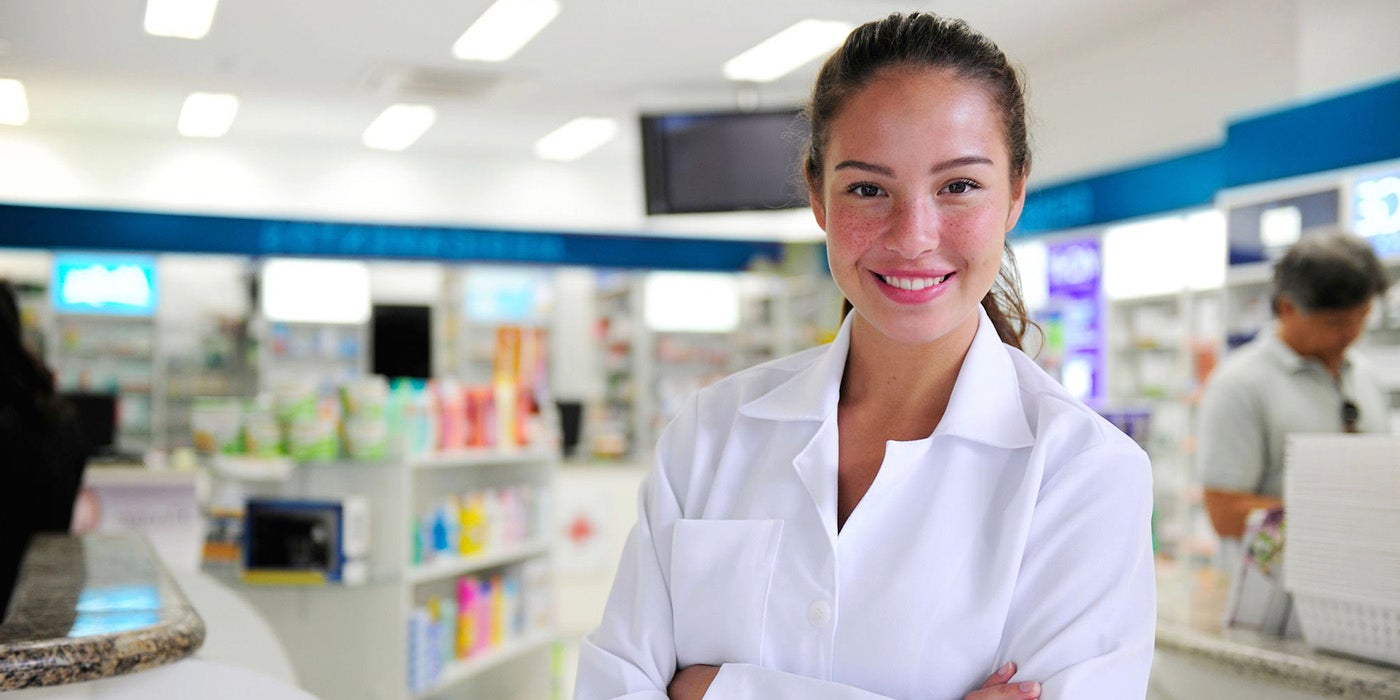 A woman in a white medical jacket in a pharmacy smiling and looking at the camera