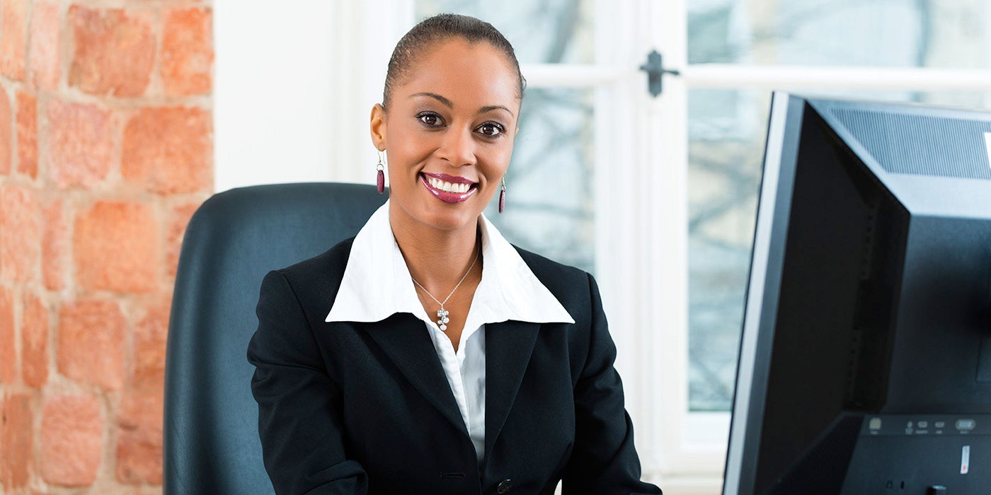 A woman dressed in a suite sitting at a desk and computer screen smiling at the camera